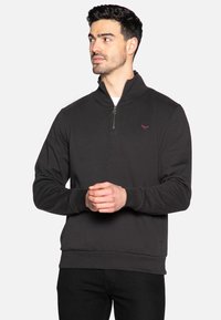 Felpa a collo alto di colore grigio scuro, con colletto a zip a quarto, polsini a coste e un piccolo logo ricamato sul petto. Tessuto morbido.