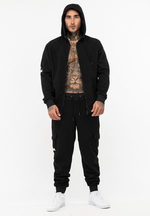 Sort zip-up hoodie med hætte, parret med sorte cargo sweatpants. Funktioner inkluderer lommer og hvide detaljer. Modellen har hvide sneakers på.