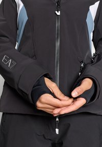 Jachetă softshell neagră, cu accente gri și alb, dotată cu fermoar frontal, manșete ajustabile și detaliu cu logo pe mânecă.