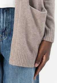 Brun stickad cardigan med ribbade manschetter och en sidoficka, bärs över högmidjade blå denim, som visar en mjuk textur.