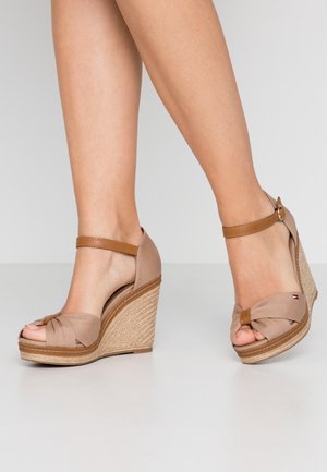 Legs wearing beige wedge sandals with ankle straps and knotted front detail, standing on a plain light floor and background.
