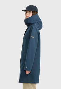 Imperméable bleu marine avec un col montant, une capuche ajustable et une manche avec logo. Texture lisse et coupe droite.