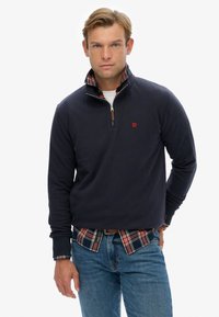 Marineblauwe pullover met een kwart rits, geribbelde manchetten en een geruite overhemd eronder. Heeft een rood logo op de borst en een zachte textuur.
