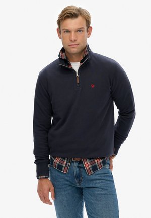 Marineblauwe pullover met een kwart rits, geribbelde manchetten en een geruite overhemd eronder. Heeft een rood logo op de borst en een zachte textuur.
