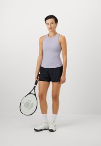 Helelilla triibuline tank top, mustad sportlikud lühikesed püksid, valged tennisetossud ja tennisereket, seismas ühtlases taustas.