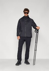 Black waterproof jacket with a front zipper and hood, worn with matching pants and shoes. Model holds black hiking poles.