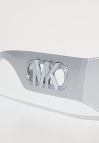 Ramele de ochelari argintii prezintă un logo proeminent "MK" cu decupaje circulare pe laturi, textură netedă și un design modern.