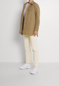 Gequilted beige overhemdjack met knoopsluiting en zak, gecombineerd met crèmekleurige ribbroeken en witte gestructureerde sneakers.