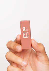 Mina leppestift i en matt ferskenrosa tube har hvit tekst som viser "503 THE LIPSTICK" på den ene siden. Hånd som holder produktet.