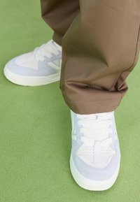 Sneaker bianche e azzurre con tomaia in suede e mesh, punta tonda, chiusura con lacci e suola testurizzata. Pantaloni marroni parzialmente visibili.