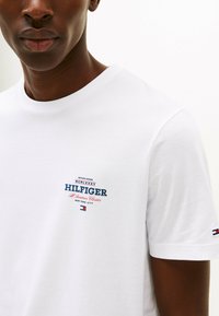 White cotton t-shirt featuring a multicolored graphic logo on the chest, with blue, red, and white text. Classic crew neck and short sleeves.