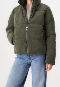 Zelená kordurová péřová bunda s vysokým límcem, zipovým uzávěrem a prošívaným vzorem, zkombinovaná s modrými džíny.