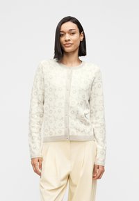 Γυναίκα που φοράει λευκό ζακέτα με λουλουδένιο σχέδιο και κουμπιά, μαζί με κρεμ ψηλόμεση παντελόνα με πιέτες, στέκεται μπροστά σε απλό φόντο.