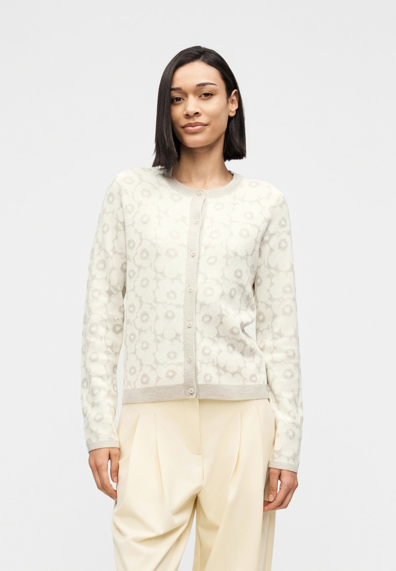 Γυναίκα που φοράει λευκό ζακέτα με λουλουδένιο σχέδιο και κουμπιά, μαζί με κρεμ ψηλόμεση παντελόνα με πιέτες, στέκεται μπροστά σε απλό φόντο.