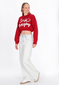 Roter, verkürzter Pullover mit weißer Aufschrift "Lächle jeden Tag", kombiniert mit hellblauen, hochgeschnittenen Jeans und beigen Sneakers.