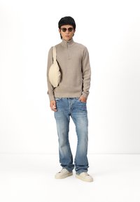 Beige ritssluiting trui, blauwe spijkerbroek, crèmekleurige sneakers en een crèmekleurige schoudertas. Model draagt zwarte zonnebril; kort, golvend haar.