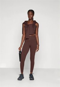 Brūns sporta apģērbs ar rakstainu topu ar kakla saiti un augsta jostasvietas legingiem. Valkāts ar melniem sporta apaviem un ūdens pudeli.