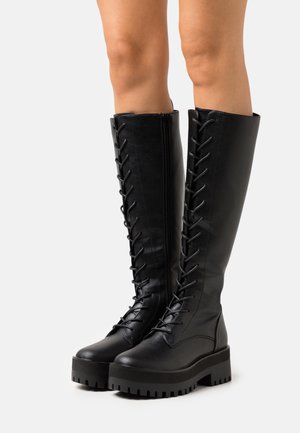 Botas negras de imitación cuero hasta la rodilla con cordones en la parte delantera, cremallera lateral y suela de plataforma gruesa con textura antideslizante.