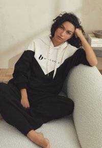 Schwarz-weiß kontrastierender Hoodie mit Logo, Kordelzügen und lockerem Schnitt. Modell sitzt auf einer strukturierten hellen Couch und trägt passende schwarze Hosen.
