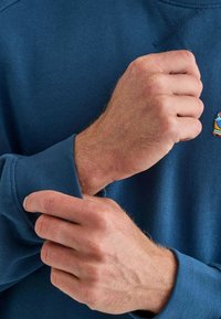 Felpa blu con polsini a costina e collo arrotondato, caratterizzata da un piccolo design ricamato vicino al petto. Il tessuto appare morbido e con una texture.