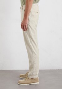 Beige cotton trousers with a straight leg, side pockets, and a single back pocket, paired with light-colored casual shoes.