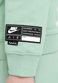 Mintgrön sweatshirt med ribbade ärmslut, med en svart etikett med vit text som inkluderar Nike-logotyp och specifikationer för idrottare.
