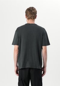 Άνδρας με κοντά ανοιχτό καφέ μαλλιά στέκεται με την πλάτη γυρισμένη, φοράει φθαρμένο μαύρο κοντομάνικο μπλουζάκι και μαύρο παντελόνι τύπου cargo μπροστά σε λευκό φόντο.