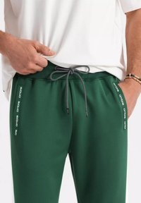 Gröna träningsoveraller med dragsko i midjan, med sidosträck i vitt. Tillverkade av ett mjukt tyg, med en avslappnad passform.