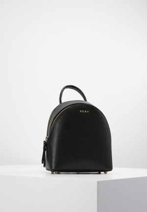 Petit sac à dos en cuir noir DKNY avec poignée supérieure, fermeture éclair dorée et logo minimaliste à l’avant, posé sur un socle blanc sur fond gris.