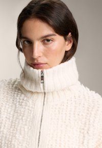 Biely pletený sveter s vysokým rebrovaným golierom a zipsovým zapínaním. Mäkká textúra s vyvýšeným vzorom po celej ploche.