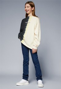 Cămașă cu mâneci lungi, colorată în nuanțe de crem și negru tie-dye, cu inscriptionarea "Sunt Liber". Poartă cu blugi albaștri și adidași albi. Textură de bumbac fin.