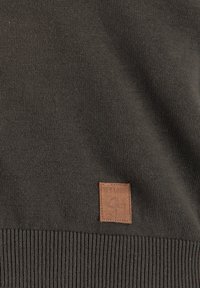 Maglione a maglia verde scuro con orlo a coste, dotato di un'etichetta rettangolare in pelle marrone con logo impresso. Texture liscia, design casual.