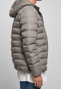 Person som bär en grå puffjacka med horisontell quiltning, huva och dragkedja framöver, över en vit skjorta och blå jeans.