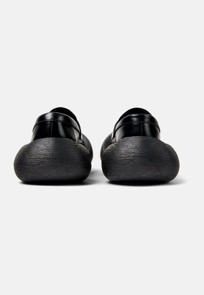 Scarpe in pelle nera con suole nere arrotondate e texture. Design elegante, hardware minimale, con una schiena aperta per indossarle facilmente.