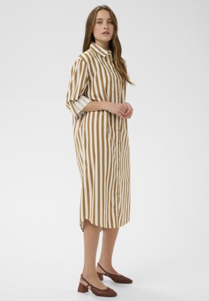 Nő hosszú, bézs és fehér, függőleges csíkos, gombos ruhában, háromnegyedes ujjúval és barna alacsony sarkú cipőben, fehér háttér előtt.