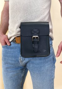 Schwarze Lederhandtasche mit einem Deckel und silberner Schnalle, am Hüftgurt getragen. Glatte Oberfläche, rechteckige Form und dekorative Stickdetails.