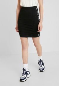 Černá klučičí sukně s přiléhavým střihem, se zvýrazněním loga na bocích. Kombinováno s bílými kotníkovými ponožkami a masivními teniskami.