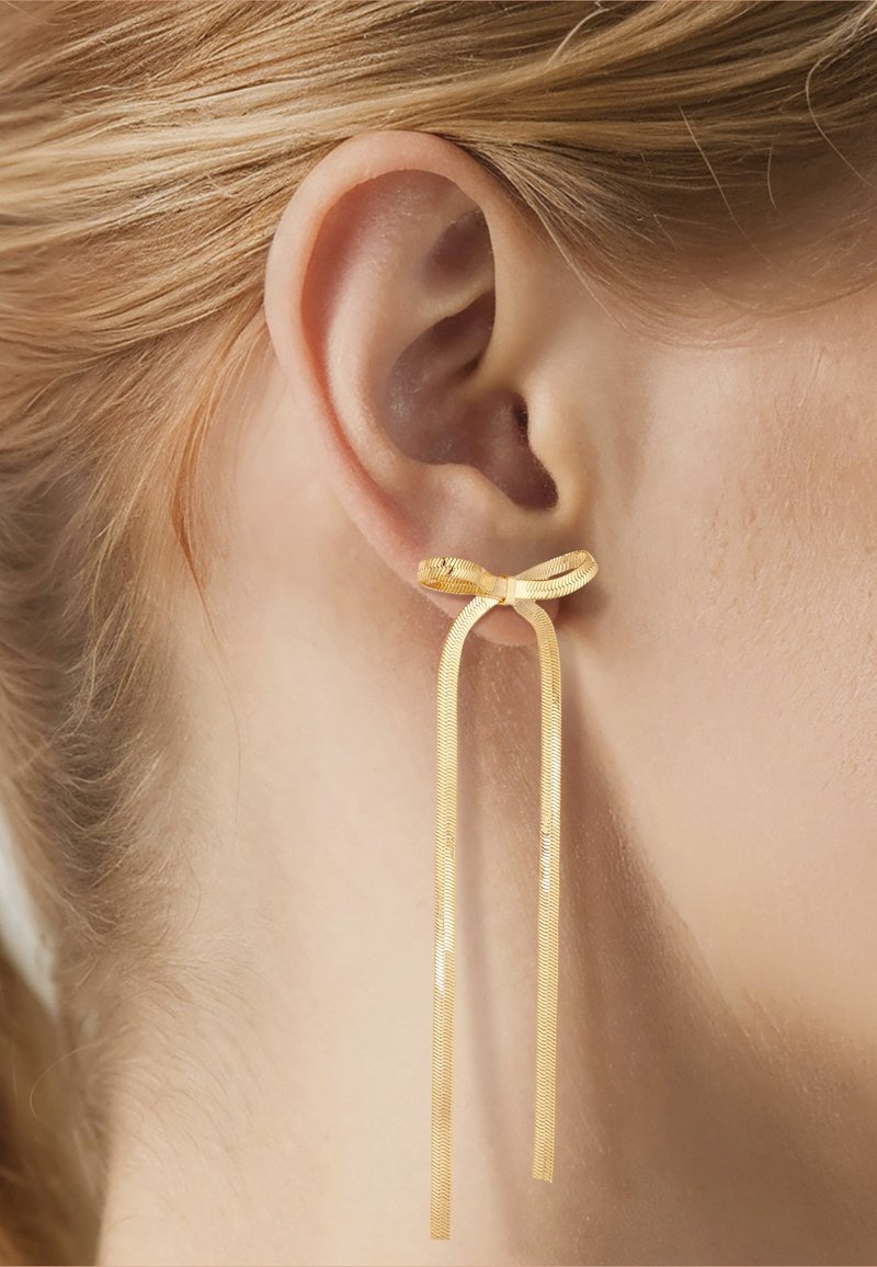 Primer plano de una oreja derecha con un pendiente dorado en forma de lazo y dos largas cadenas doradas colgando del lazo.