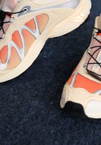 Chaussures de sport en crème, orange et argent, composées de matériaux en maille et synthétiques. Présentent des côtés texturés, des lacets noirs et une semelle robuste.