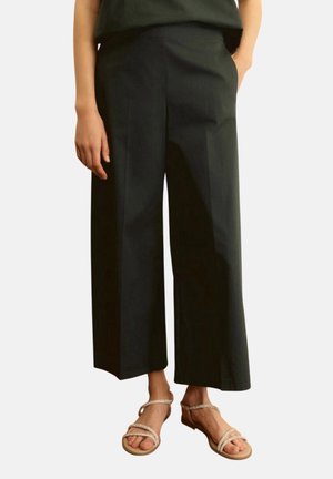 Pantaloni neri a gamba larga e corta indossati con sandali beige con tacco basso e cinturini, una mano in tasca, in piedi su sfondo uniforme.