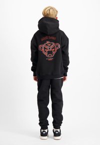 Zwarte hoodie met een grafisch ontwerp van een gestileerd berenhoofd, gecombineerd met zwarte joggingsbroek en zwarte sneakers met witte accenten.