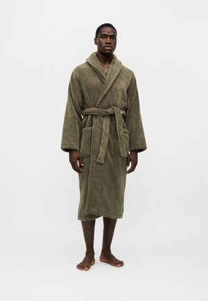 Homme debout pieds nus portant un peignoir en éponge vert olive ceinturé avec poches avant, sur un fond clair uni.