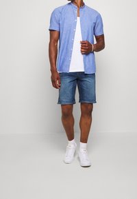 Hombre vestido con una camisa de botones de manga corta azul clara sobre una camiseta blanca, pantalones cortos de mezclilla, calcetines blancos y zapatillas blancas, de pie contra un fondo liso.