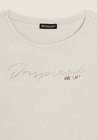 Camiseta de color gris claro con escote redondo, que presenta "Inspired HEART" en rhinestones y detalles metálicos, material de algodón suave.