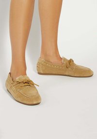 Tan suede loafers with a lace tie, gold studs along the upper edge, and a soft fluffy lining. Flat soles for comfort.