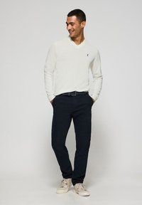 Weißer V-Ausschnitt-Pullover mit gerippten Bündchen, marineblaue Chinos und helle Sneaker. Enthält ein kleines Logo auf der linken Brust. Lässige Passform und strukturierter Stoff.