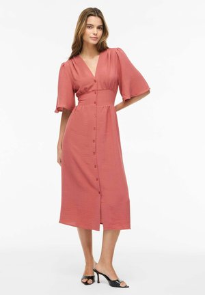 Kvinde står iført en tekstureret, knap-ned, støvet lyserød midi-kjole med albuelange ærmer og sorte højhælede sandaler.