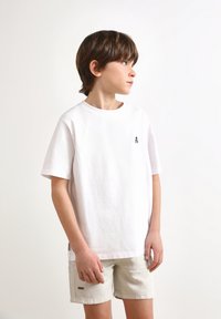 Camiseta blanca de algodón con mangas cortas, que presenta un pequeño logo negro en el pecho. Combinada con pantalones cortos beige claros con un detalle sutil.