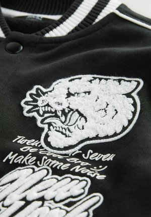 Veste varsity noire avec un design brodé texturé blanc représentant la tête d'un animal grognant et du texte stylisé en blanc. Col rayé.