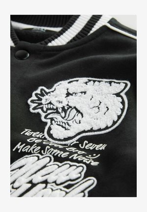 Crna jakna varsity s teksturiranim bijelim vezom koji prikazuje njušku zveri koja reži, te bijelim stiliziranim tekstom. Prugasti ovratnik.